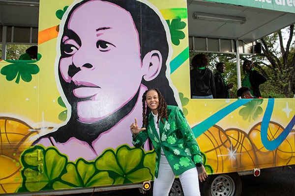 Seimone Augustus in a green sequined shamrock jacket smiling and making a LSU sign in front of a colorful mural with a large portrait of herself with basketballs and clover leaves.