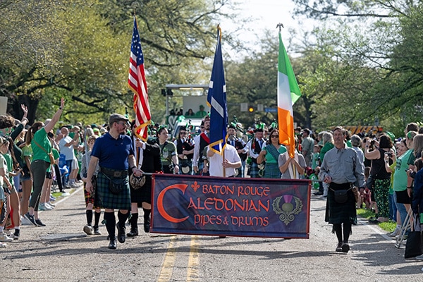 A vibrant parade with participants marching down a tree-lined street, wearing green and carrying the American and Irish flags, followed by a banner for the Baton Rouge Caledonian Pipes & Drums, suggesting a