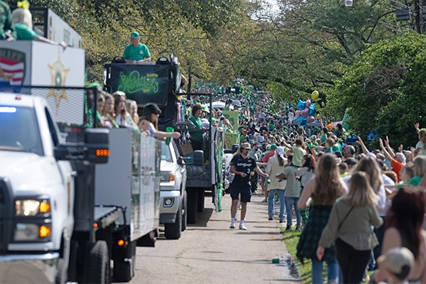 A 2024 festive St. Patrick's Day parade in full swing with people dressed in green, floats, and a lively crowd celebrating along the route.
