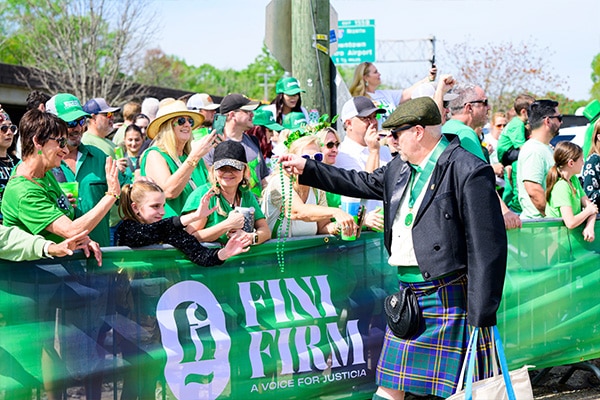 A lively St. Patrick's Day parade scene with a man in a kilt and a beret interacting with the crowd, as bystanders dressed in green with festive accessories engage and reach out for beads
