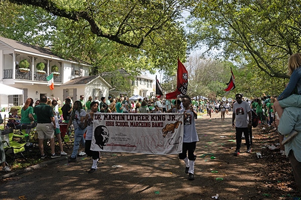 A lively parade with spectators lining the street as members of the Dr. Martin Luther King Jr. High School marching band, dressed in Wearin' of the Green attire, march proudly, carrying banners and