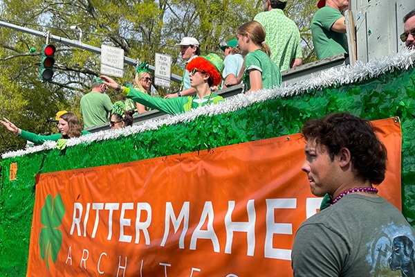 A vibrant street parade with people wearing green, celebrating the Wearin' of the Green on a float decorated with the words 'Ritter Maher Architects,' as a man watches the festivities.