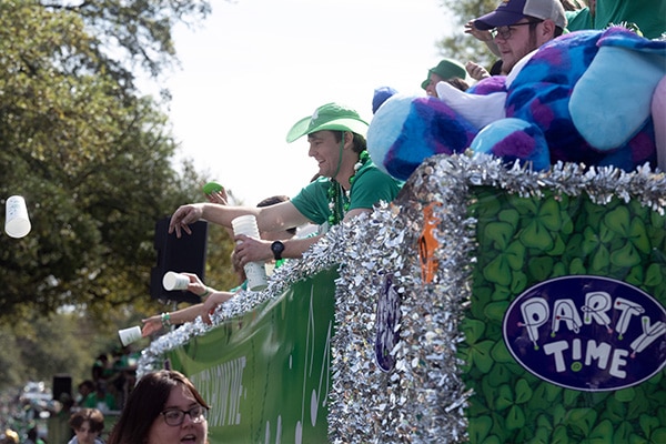 A festive parade float with people dressed in green for the 2024 Wearin' of the Green, throwing beads and enjoying the celebration.