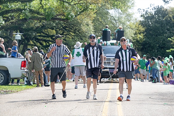 Baton Rouge Ancient Athletes a popular group dressed as referees, one carrying a whistle, lead the Wearin' of the Green parade on a sunny day, with onlookers lining the route and a float visible in the