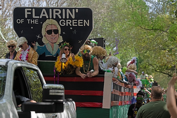 A vibrant and festive parade float with the theme "Wearin' of the Green," featuring individuals in costumes and sunglasses, tossing beads to the crowd, set against a backdrop of lush greenery in