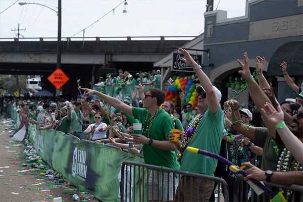 A lively crowd dressed in green with beads and glasses celebrates the 2024 Wearin' of the Green at an outdoor event, possibly a parade, with a barricade in the foreground and festive debris