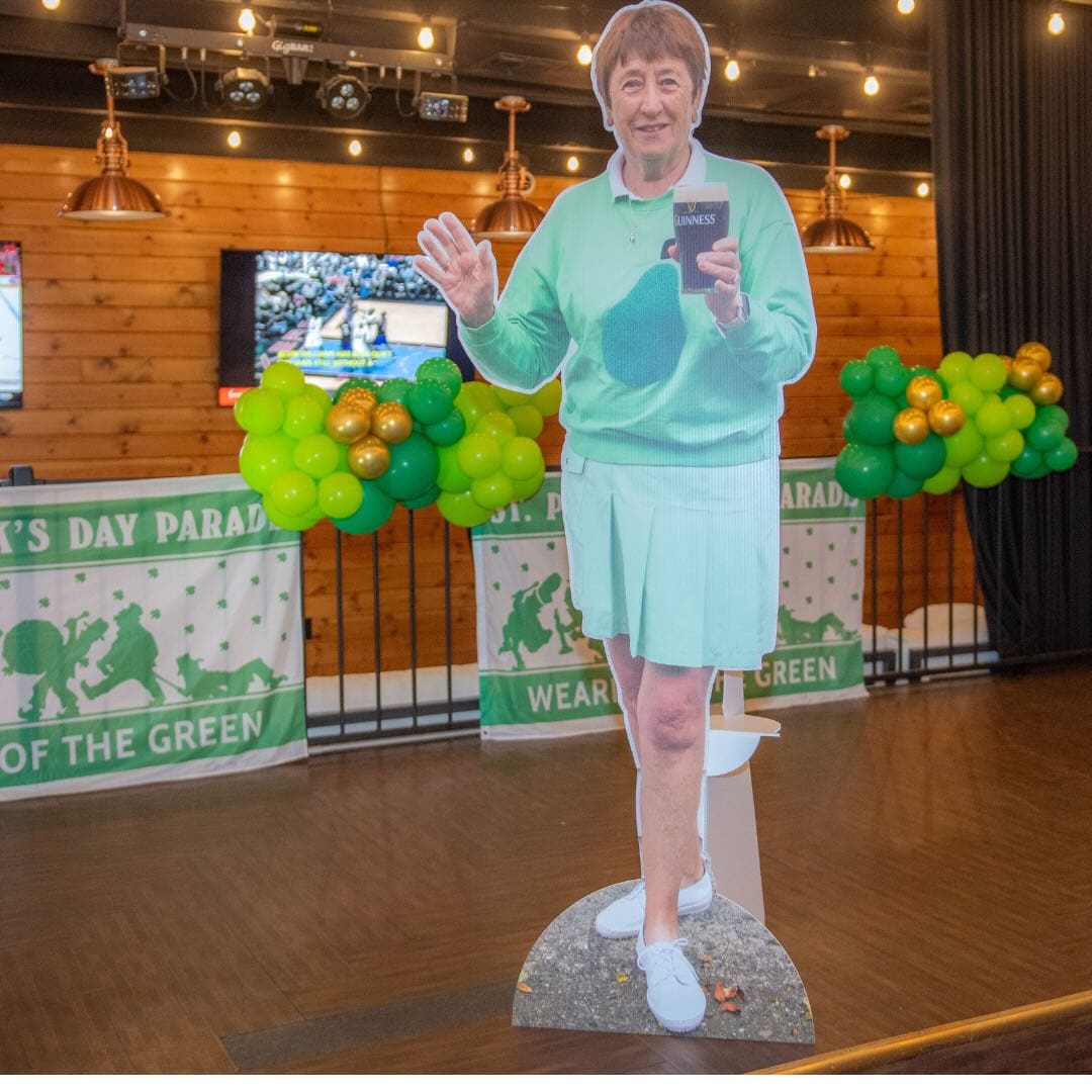 Cardboard cutout of a person wearing a light green outfit, holding a book, and waving. Green and gold balloons and St. Patrick's Day banners in the background add to the festive spirit reminiscent of Noreen Coffey's lively celebrations.