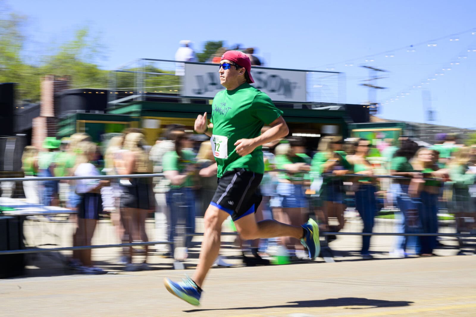 A man in a green shirt and red headband runs in the Shamrock Run, with a blurred crowd in green shirts watching in the background.