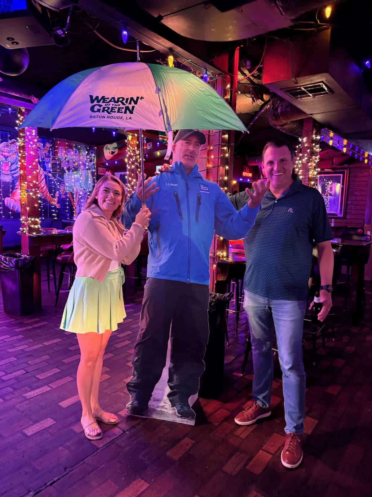 Two people pose with a Grand Marshals life-size cardboard cutout, surrounded by colorful string lights in a lively bar.